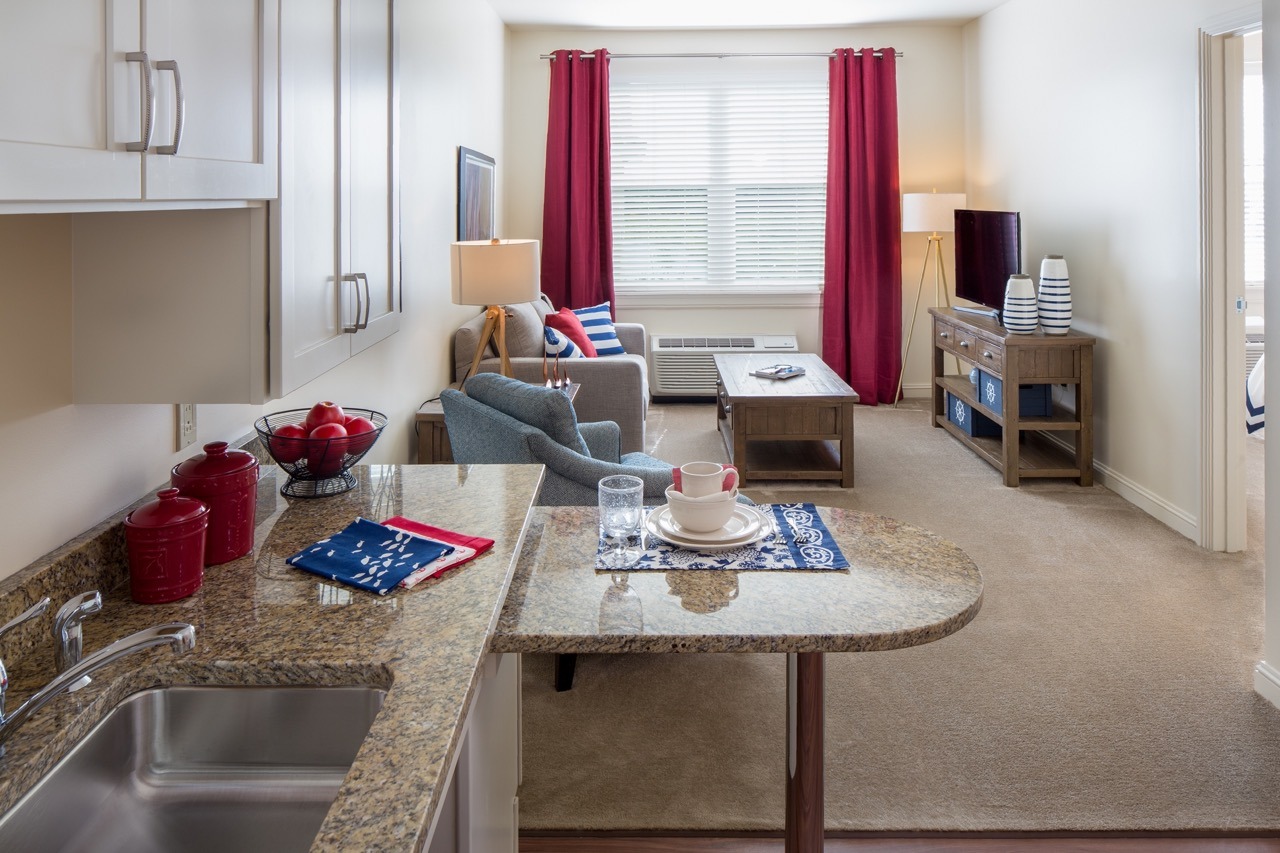A view of the living room from the kitchen, showing a couch, a blue sofa chair, a coffee table, a TV on a TV stand, and a window with open red curtains at The Summit of Lakewood Ranch.